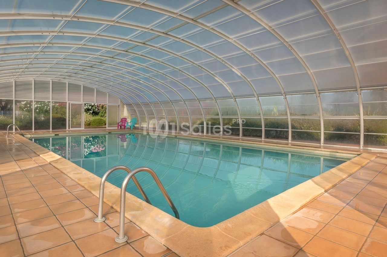 Piscine intérieure couverte par une structure transparente, entourée de carrelage beige et de quelques chaises colorées.