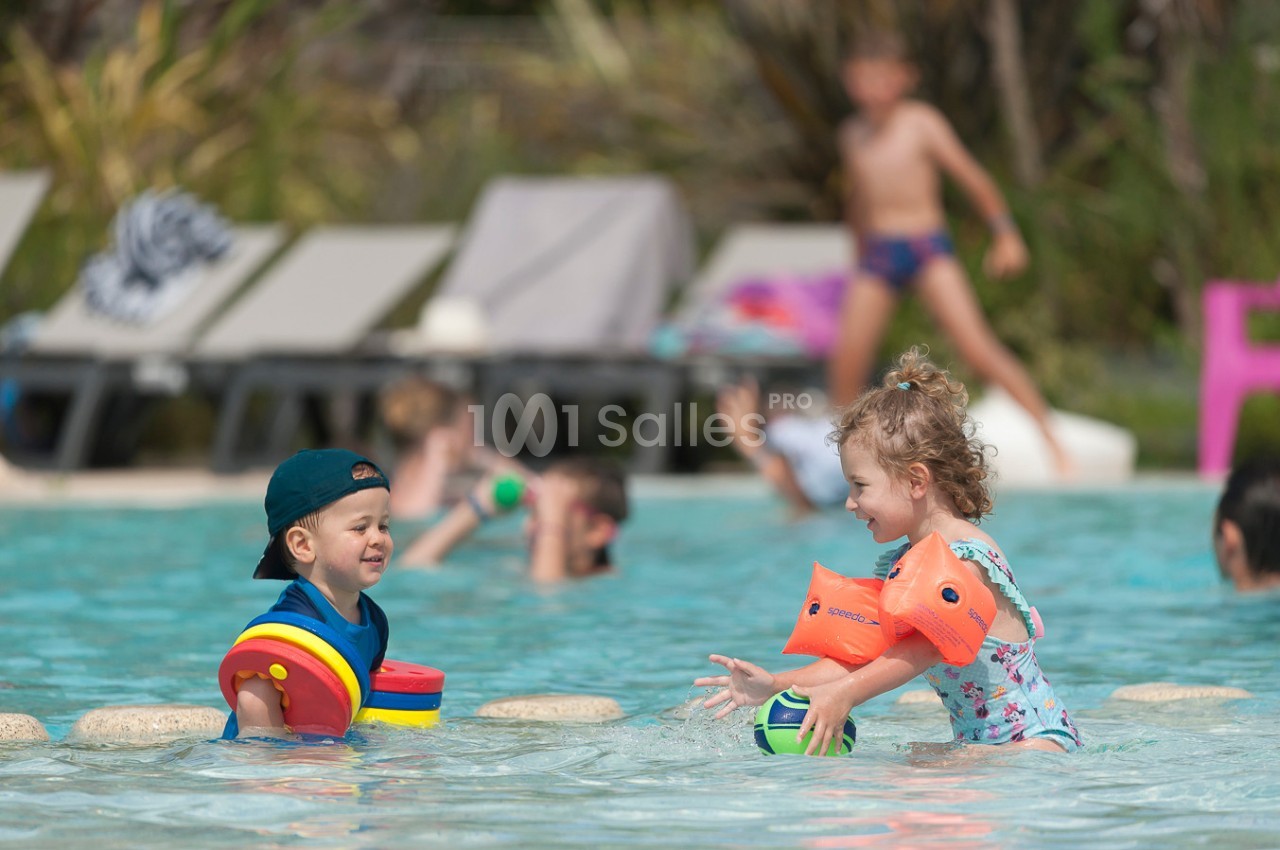 Deux jeunes enfants jouent avec des jouets flottants dans une piscine peu profonde, entourés d'autres baigneurs.