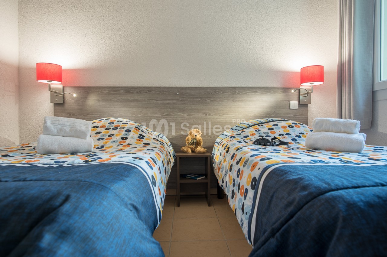 Deux lits simples avec couvre-lits colorés, serviettes pliées et une peluche au centre sur une table de chevet.