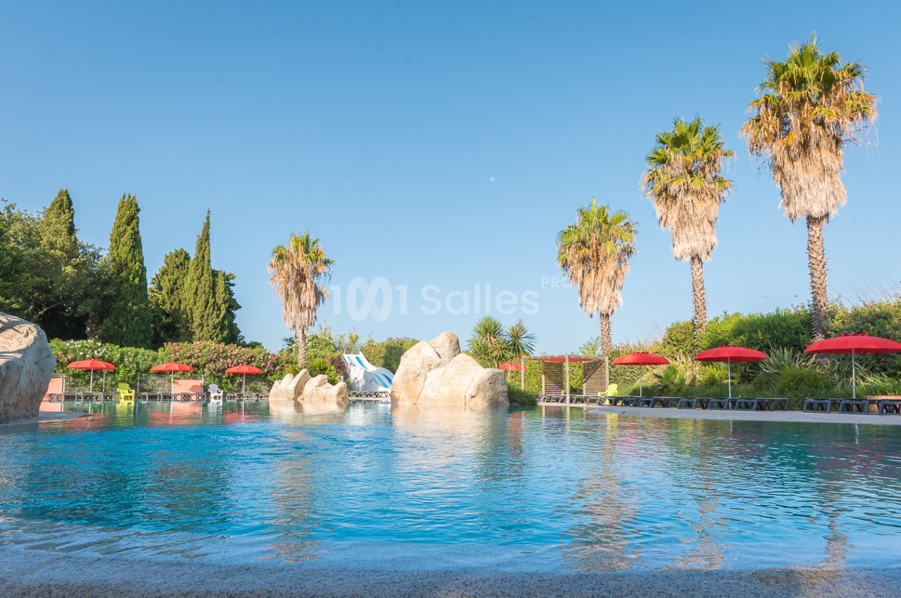 Piscine extérieure entourée de palmiers, parasols rouges et végétation sous un ciel bleu clair.