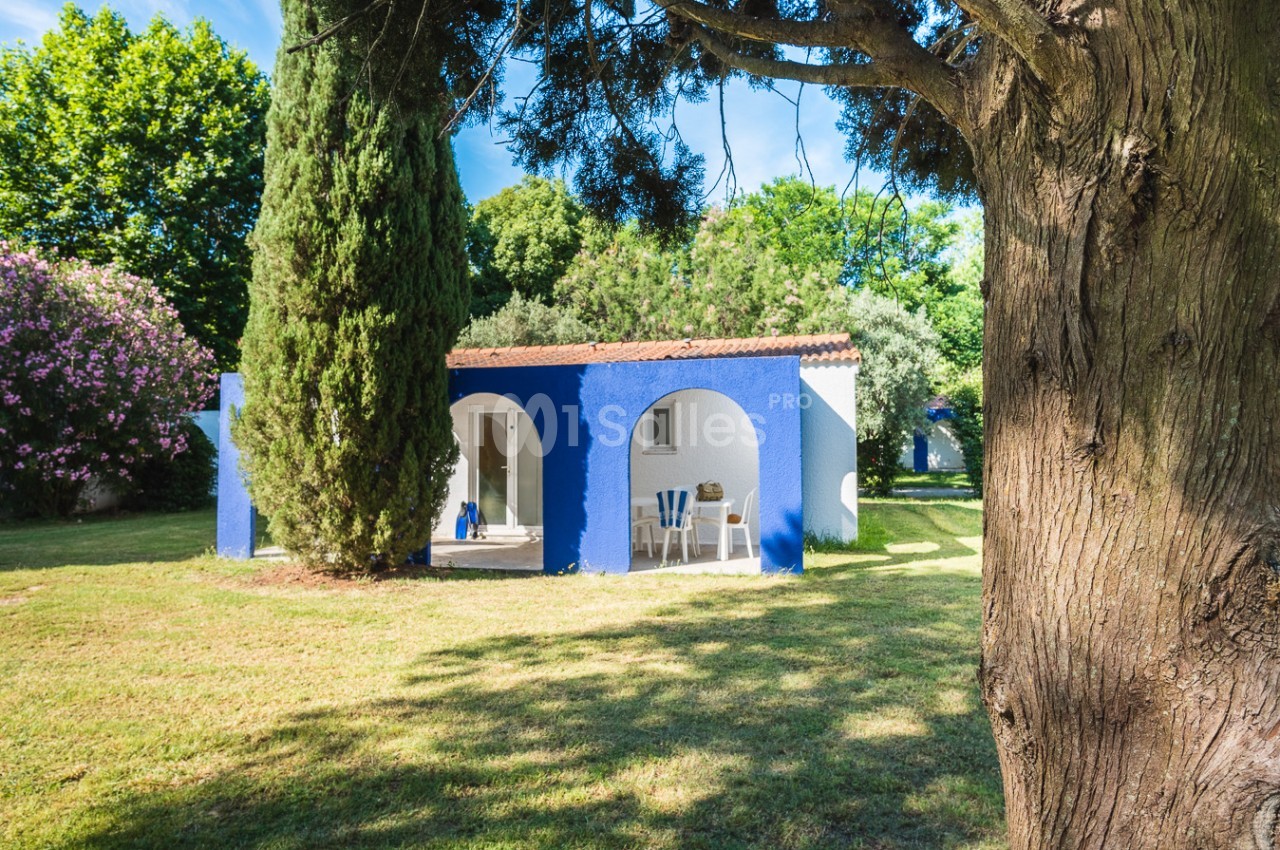 Petite maison blanche et bleue entourée d'arbres et d'arbustes, située dans un jardin ensoleillé.