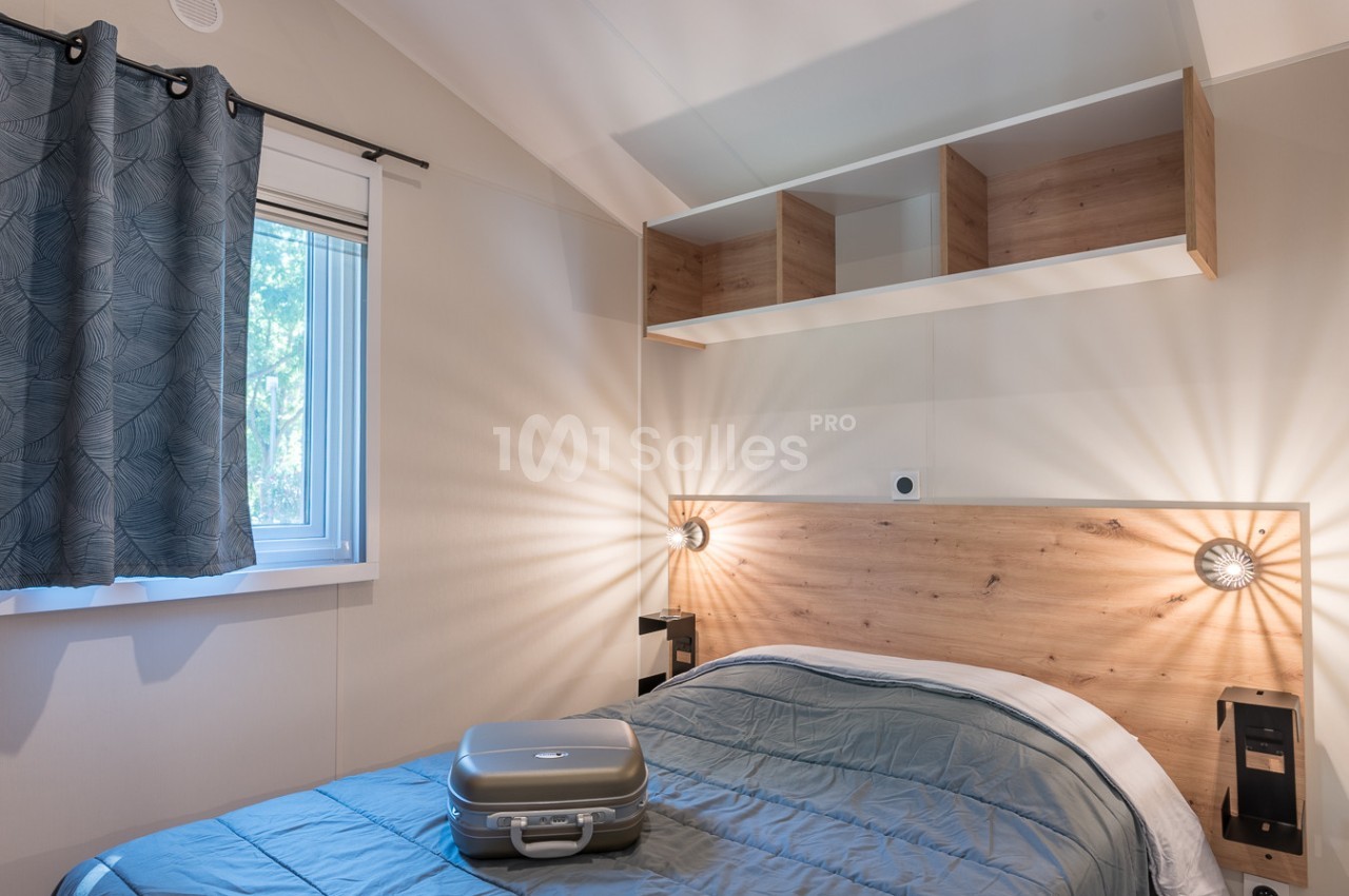 Chambre lumineuse avec un lit double, une valise posée dessus, une fenêtre avec rideaux et des étagères murales en bois.