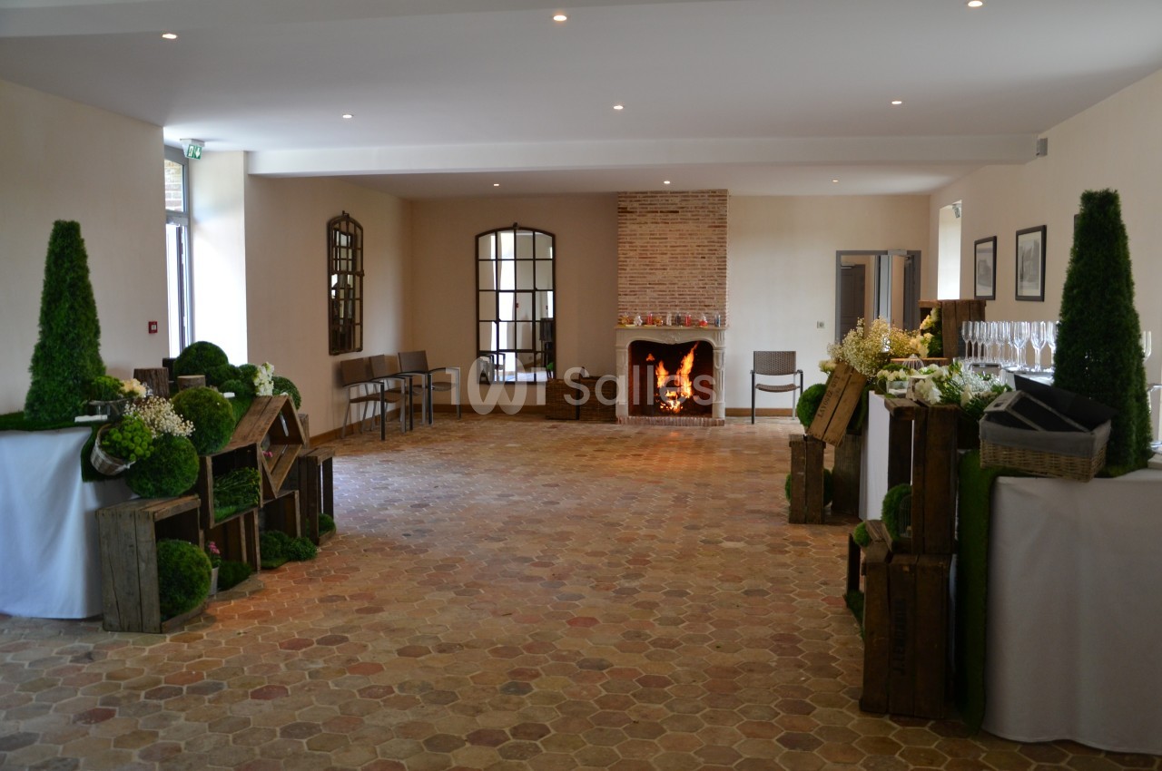 Salle lumineuse avec sol en carreaux de terre cuite, cheminée allumée et décorations végétales sur des caisses en bois.