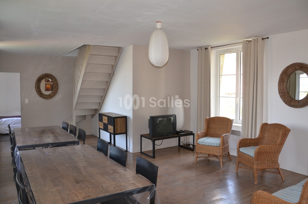 Salle de séjour lumineuse avec table en bois, chaises noires, fauteuils en rotin, télévision et escalier menant à l'étage.
