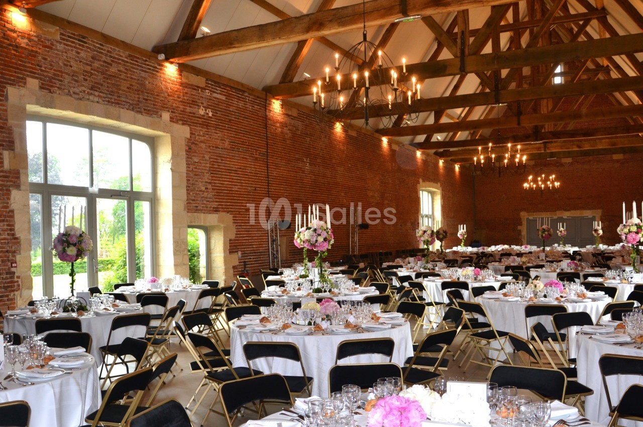 Salle de réception en briques avec poutres apparentes, tables rondes dressées et décorations florales élégantes.
