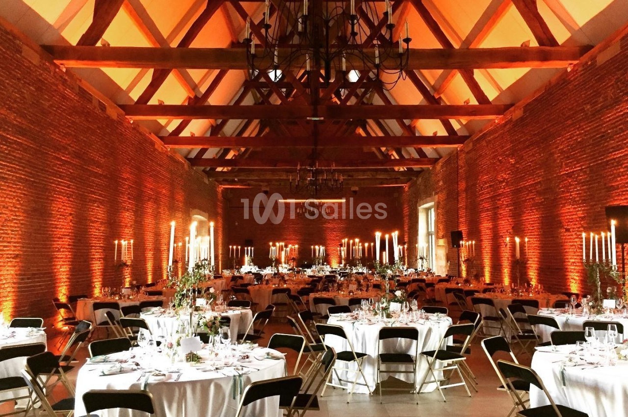 Salle de réception en briques avec charpente apparente, tables rondes dressées et éclairage chaleureux.