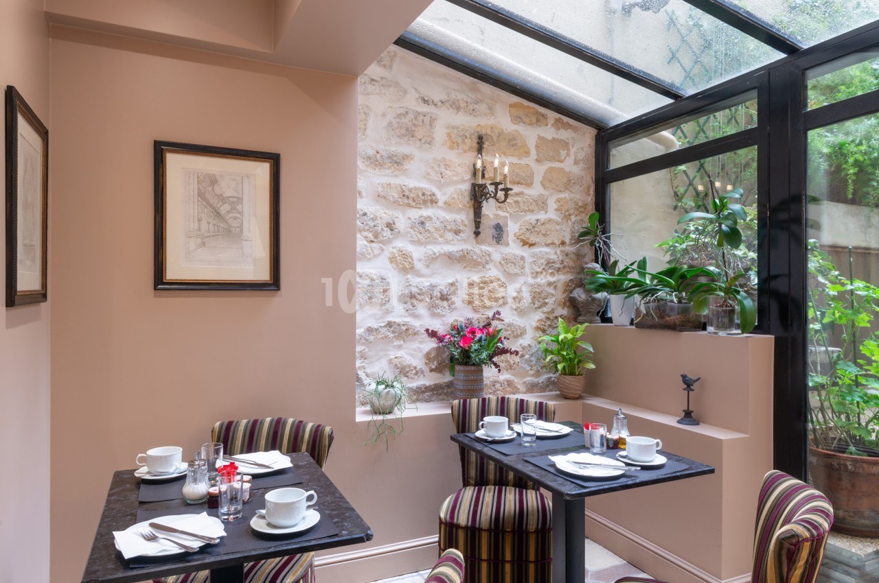 Salle de restaurant lumineuse avec murs en pierre, verrière, plantes et tables dressées pour le petit-déjeuner.