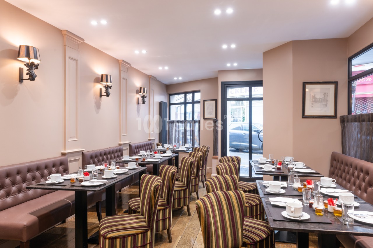 Salle de restaurant lumineuse avec tables dressées, chaises rayées et murs beige, vue sur des fenêtres donnant sur la rue.