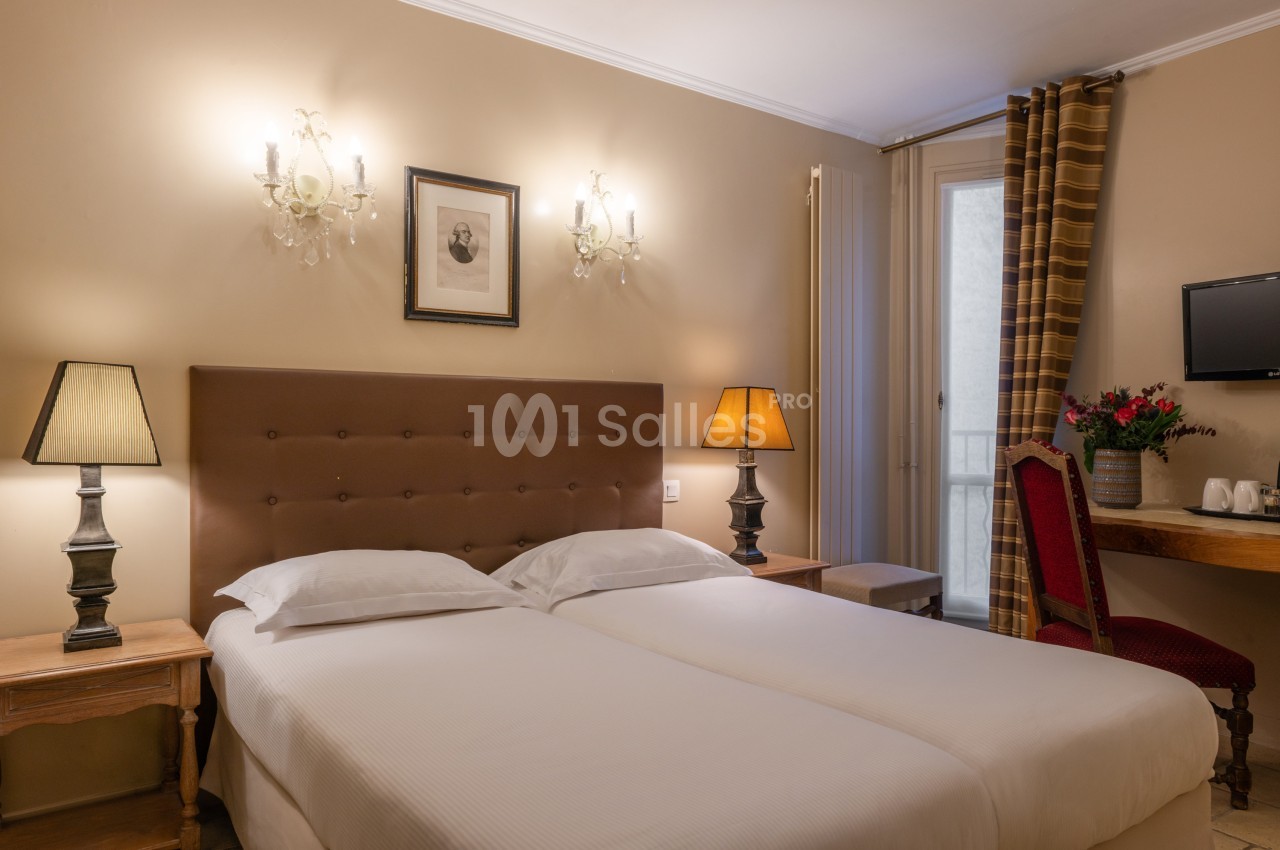 Chambre avec deux lits simples, éclairée par des lampes de chevet, avec un bureau, une chaise et une télévision murale.