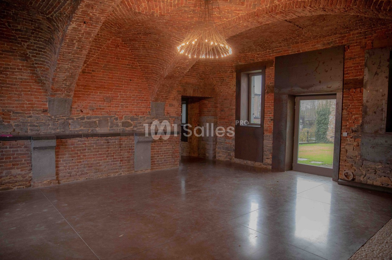 Salle voûtée en briques avec un sol en béton poli, éclairée par un lustre moderne et donnant sur un jardin.
