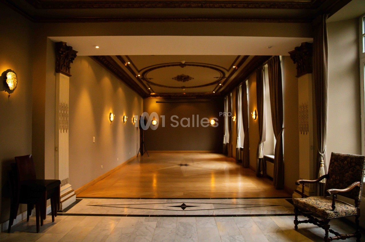 Salle élégante avec parquet, moulures au plafond, murs beige, appliques murales et grandes fenêtres avec rideaux.