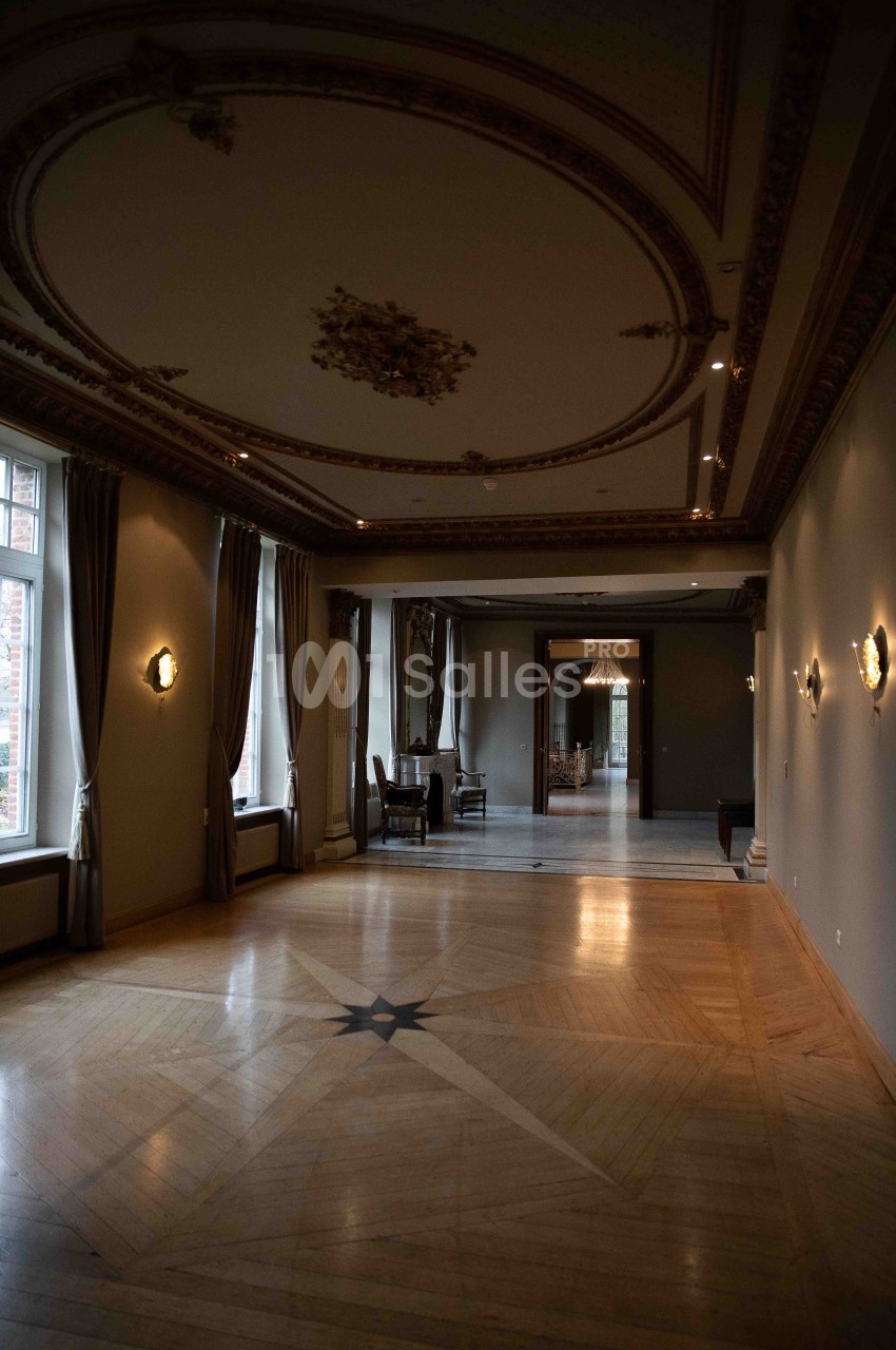 Salle vide avec parquet en bois, moulures au plafond, grandes fenêtres et rideaux, éclairée par des appliques murales.