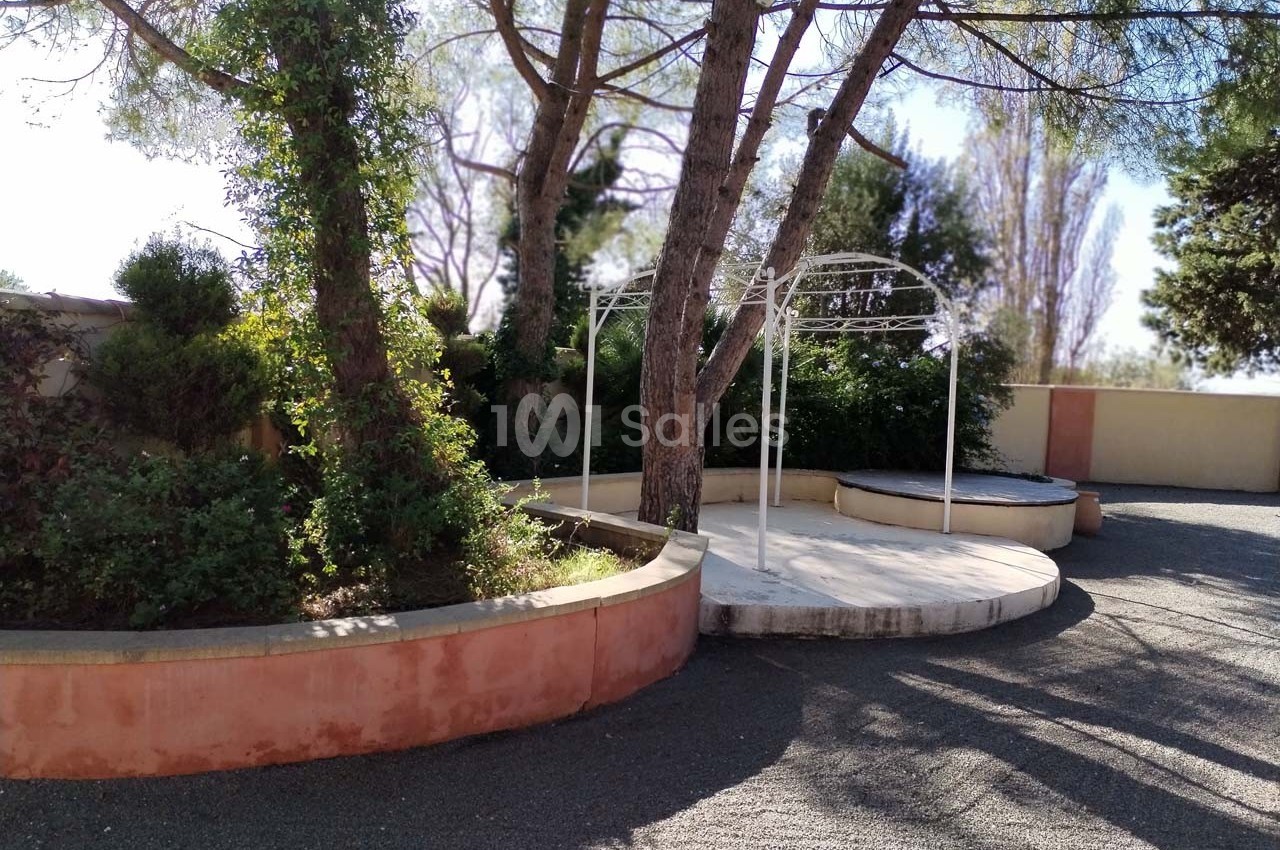 Terrasse extérieure avec pergola blanche, entourée de végétation et de murs en pierre sous des arbres.