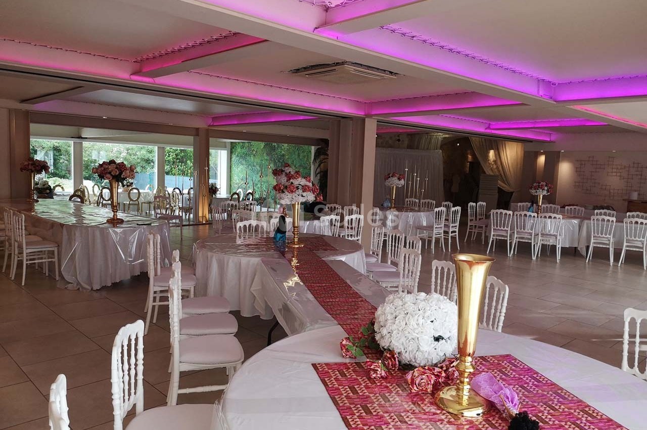 Salle de réception décorée avec des tables rondes, nappes blanches, centres de table fleuris et éclairage rose au plafond.