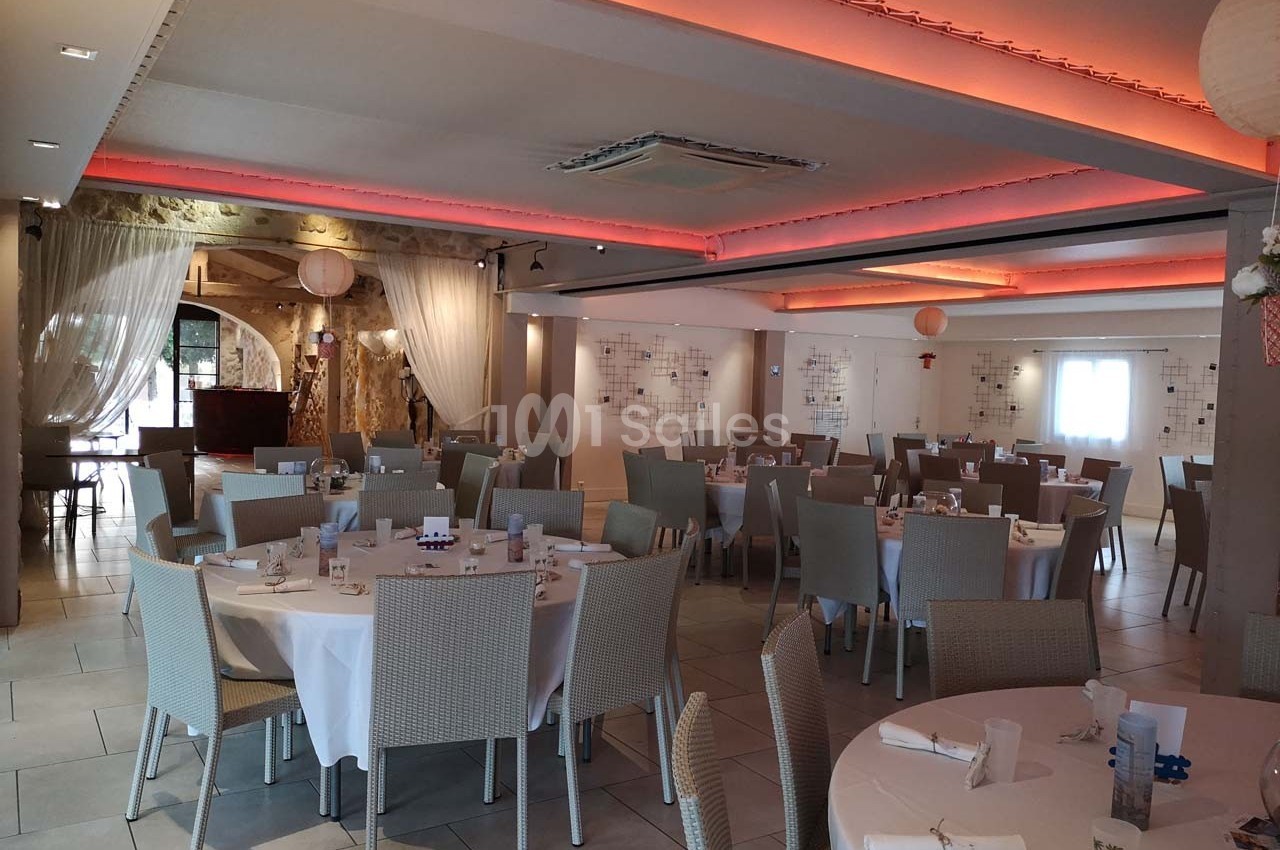 Salle de réception lumineuse avec tables rondes dressées, chaises en osier clair et éclairage tamisé au plafond.