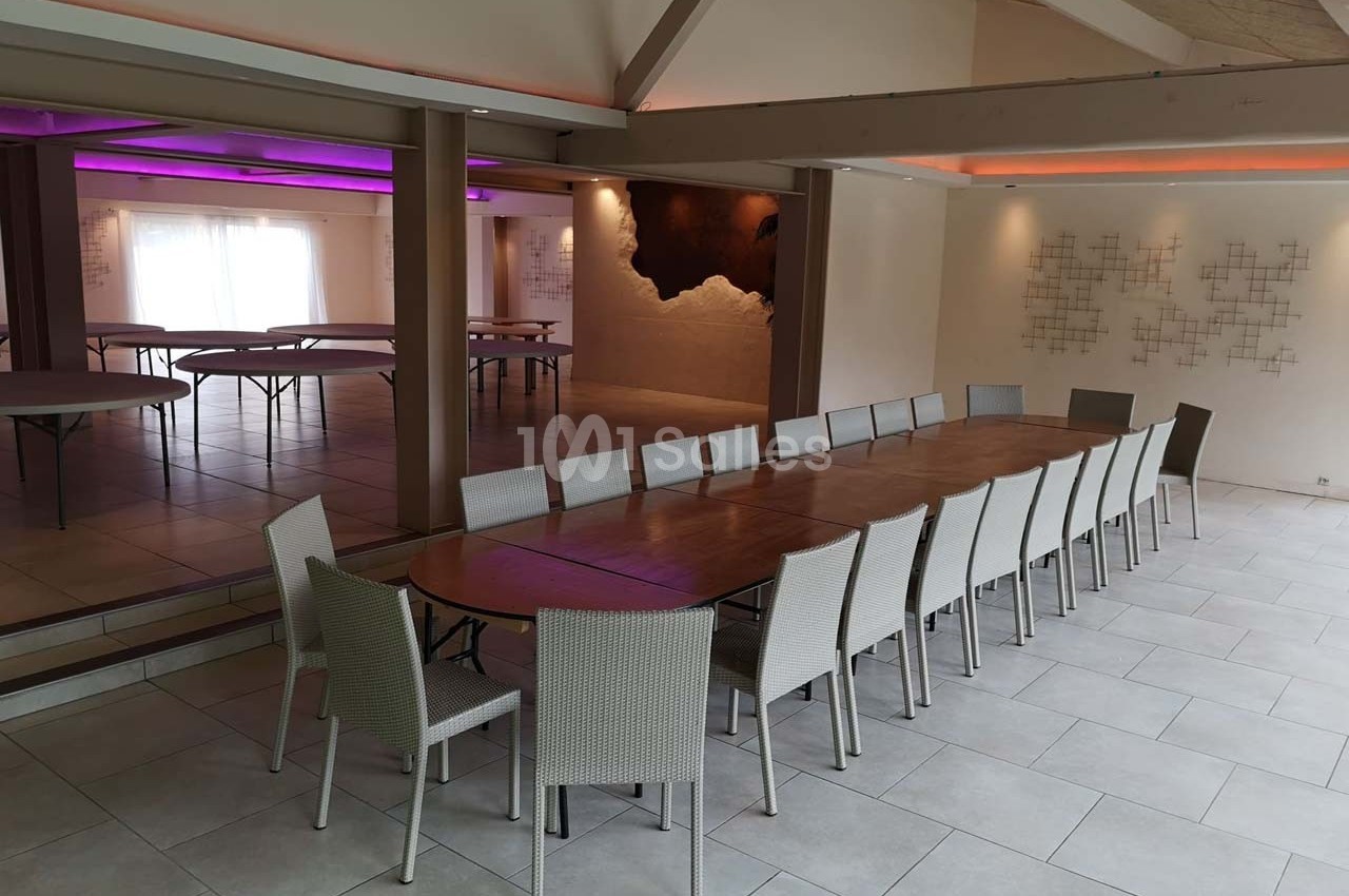 Grande salle lumineuse avec une longue table en bois entourée de chaises, murs décorés et éclairage coloré en arrière-plan.