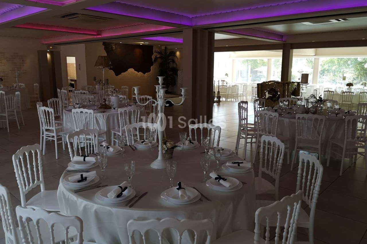 Salle de réception décorée avec des tables rondes, nappes blanches, chaises élégantes et éclairage tamisé.