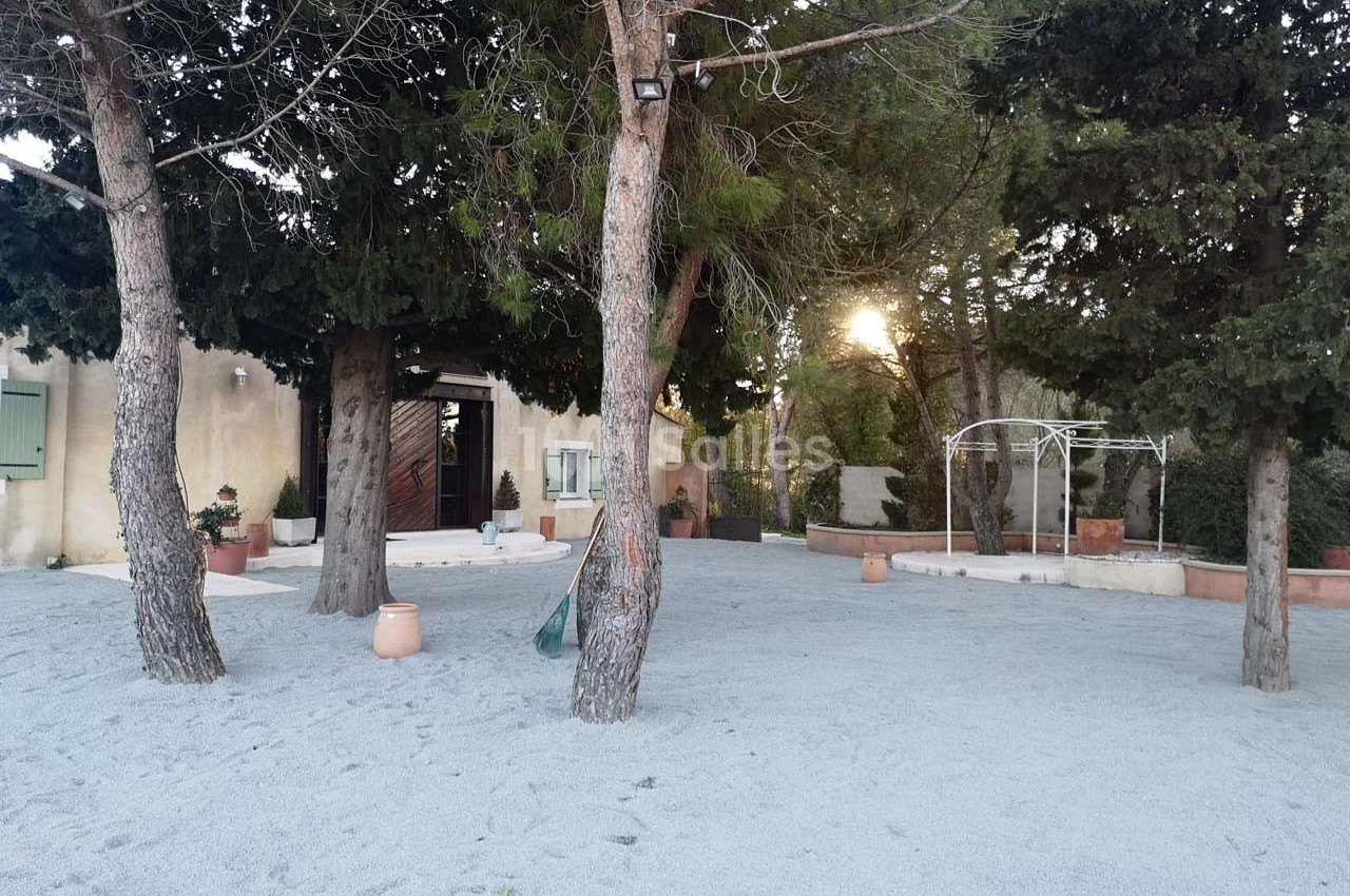 Cour extérieure avec sol en gravier, arbres, maison beige en arrière-plan et structure métallique blanche à droite.