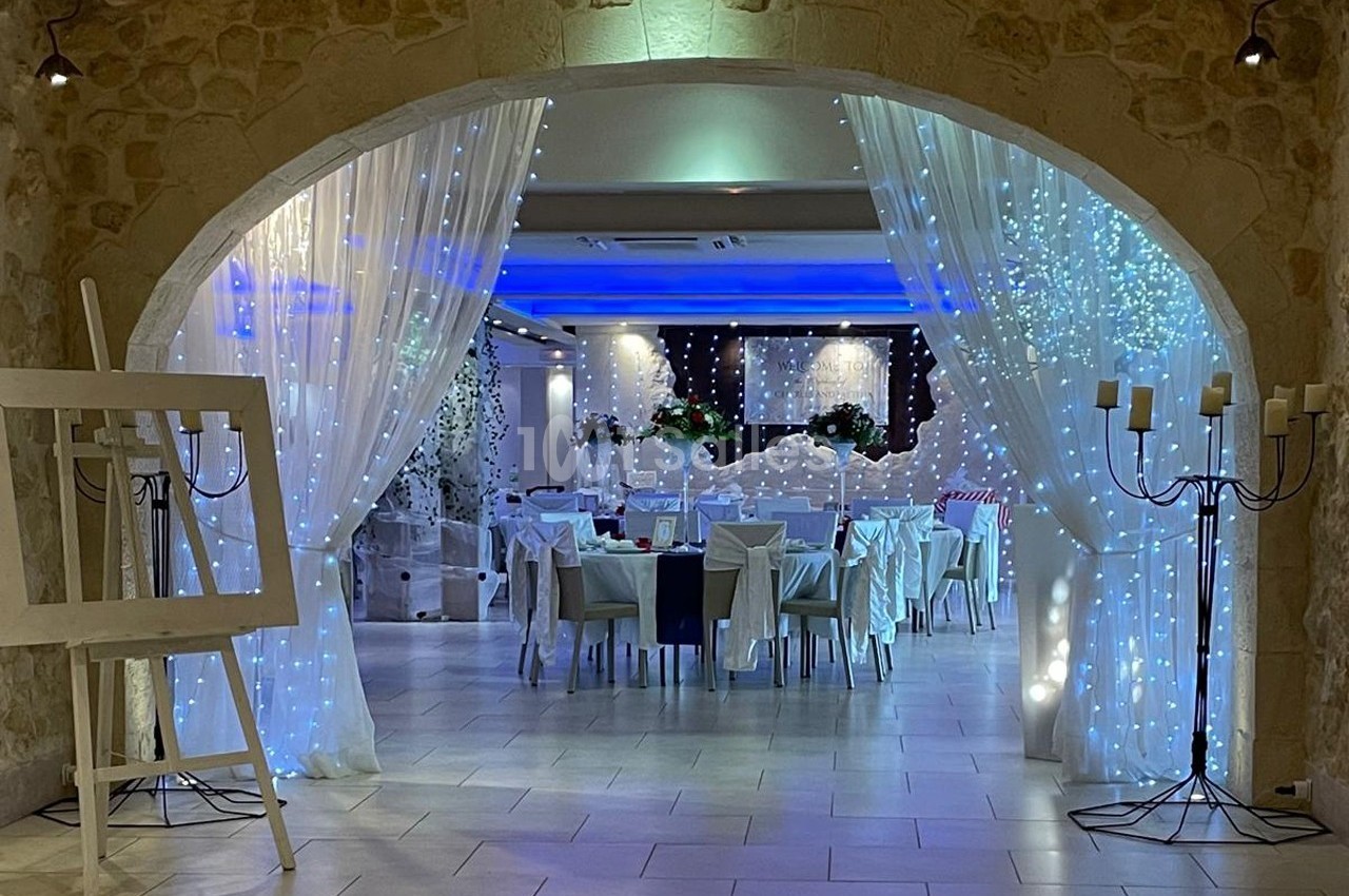 Salle de réception décorée avec des rideaux blancs, guirlandes lumineuses et tables dressées pour un événement.