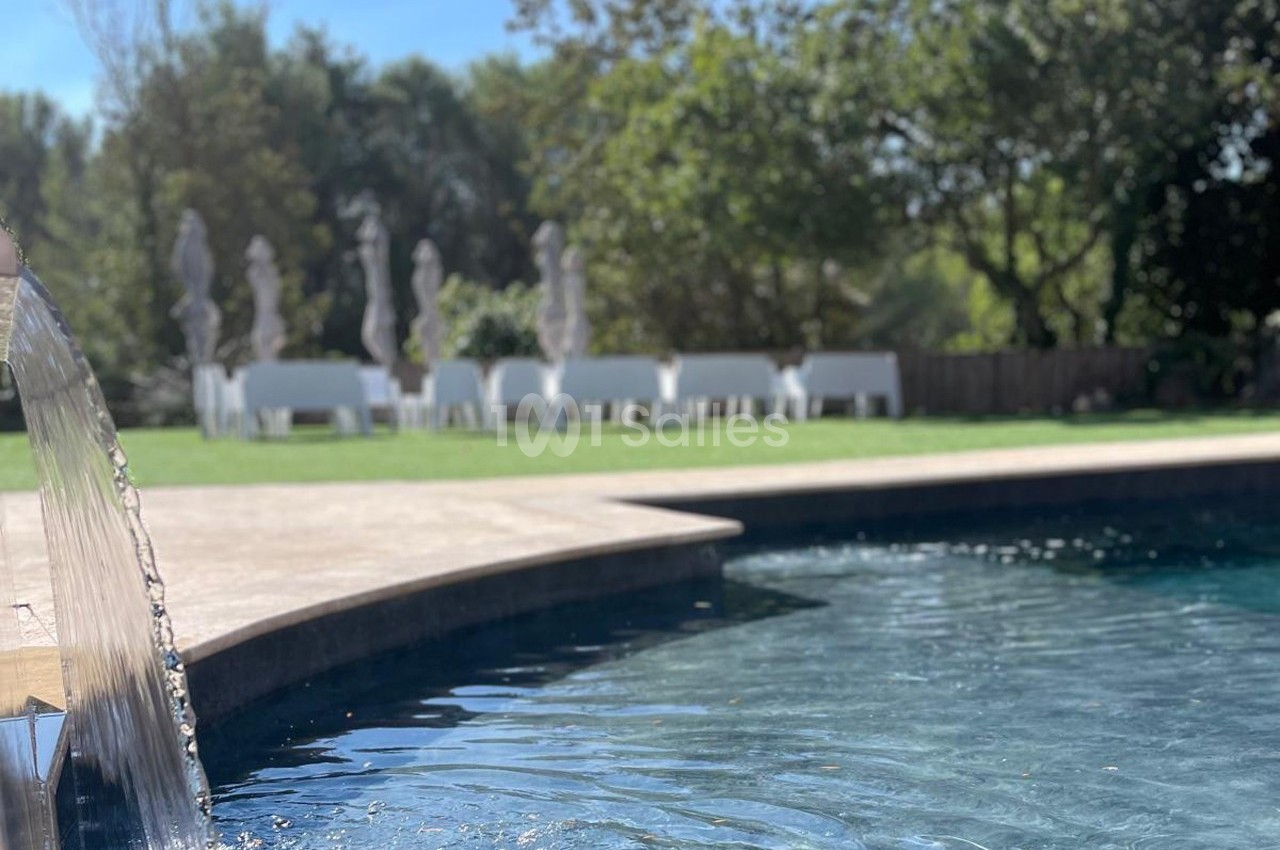 Eau d'une piscine avec une cascade au premier plan, pelouse et tables blanches sous des parasols en arrière-plan.
