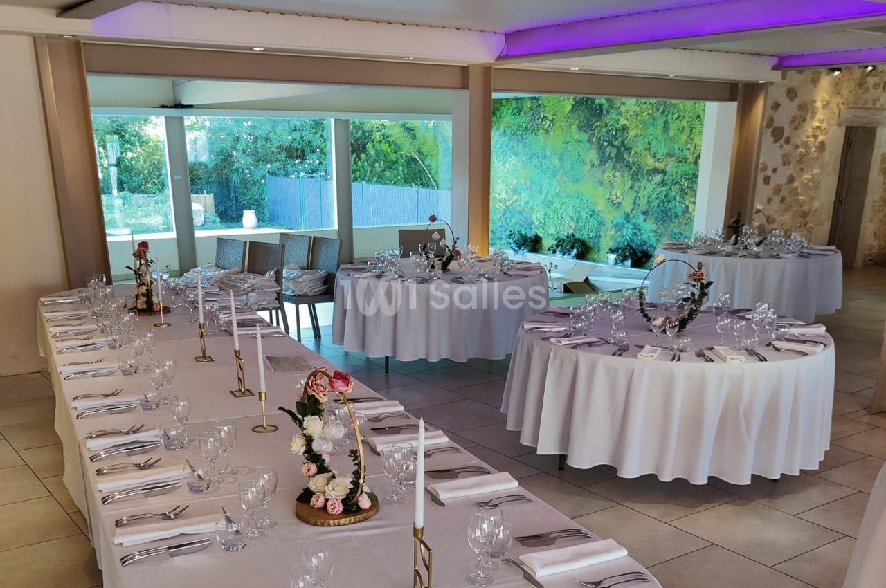 Salle de réception décorée avec des tables dressées, nappes blanches, chandeliers et vue sur un jardin verdoyant.