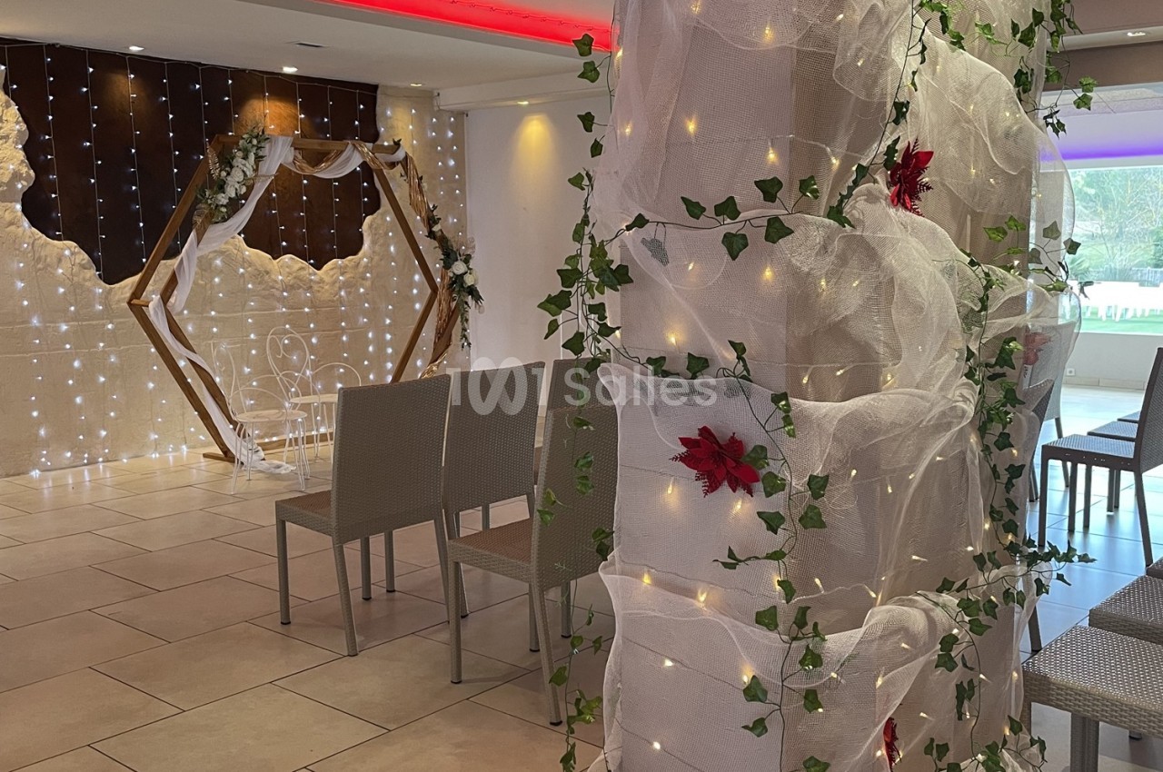 Salle décorée pour une cérémonie avec arche en bois, chaises alignées et guirlandes lumineuses sur des voilages.