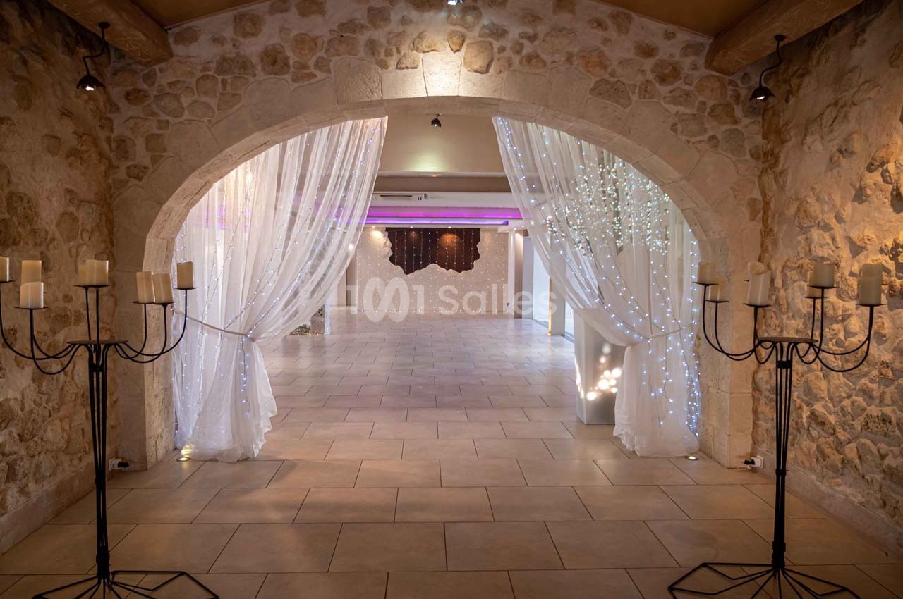 Salle en pierre avec arche, rideaux blancs lumineux et chandeliers, offrant une ambiance élégante et chaleureuse.