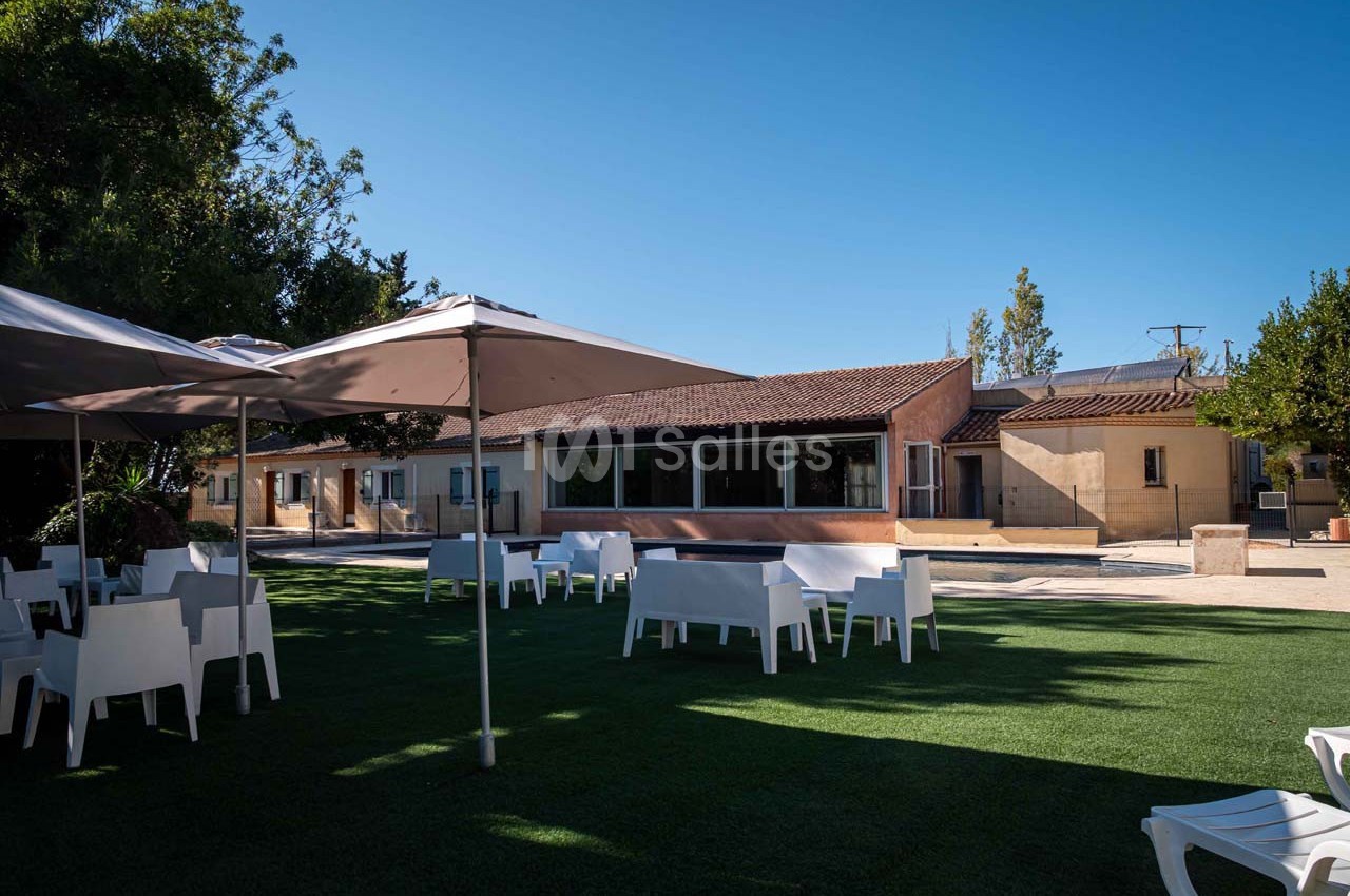 Espace extérieur avec pelouse, mobilier blanc et parasols, devant un bâtiment de style méditerranéen sous un ciel dégagé.