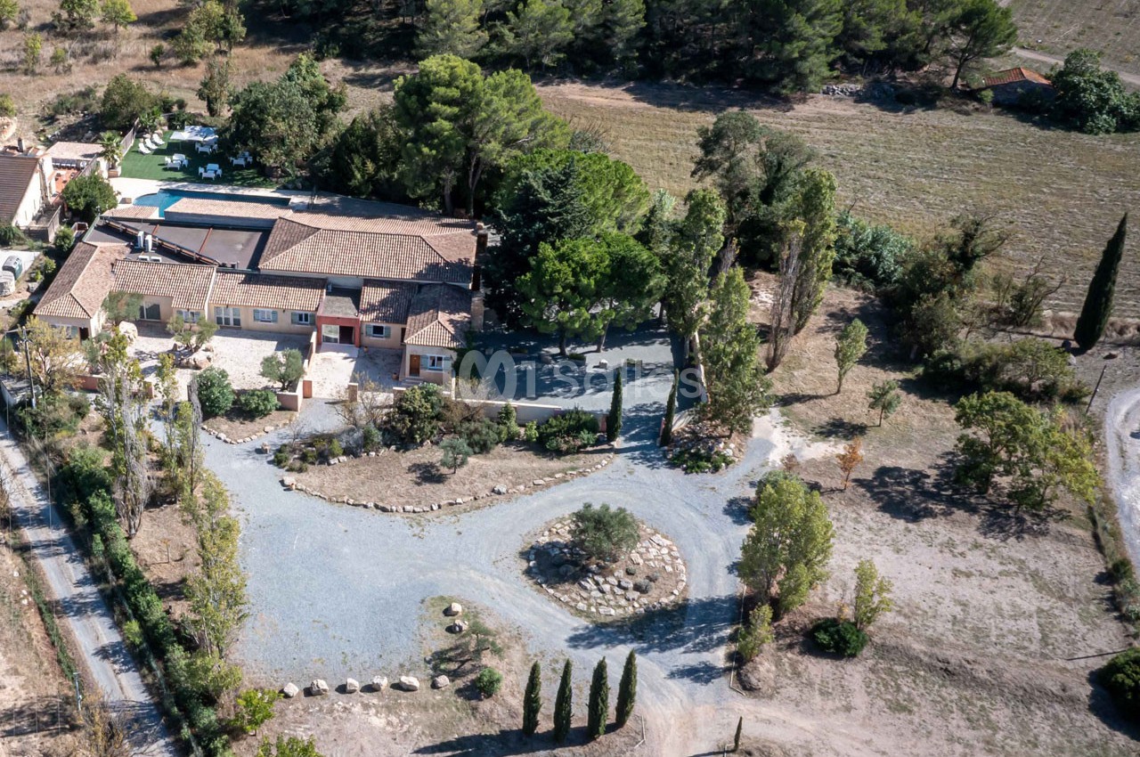 Vue aérienne d'une propriété entourée de végétation, avec bâtiments, piscine et allées gravillonnées.