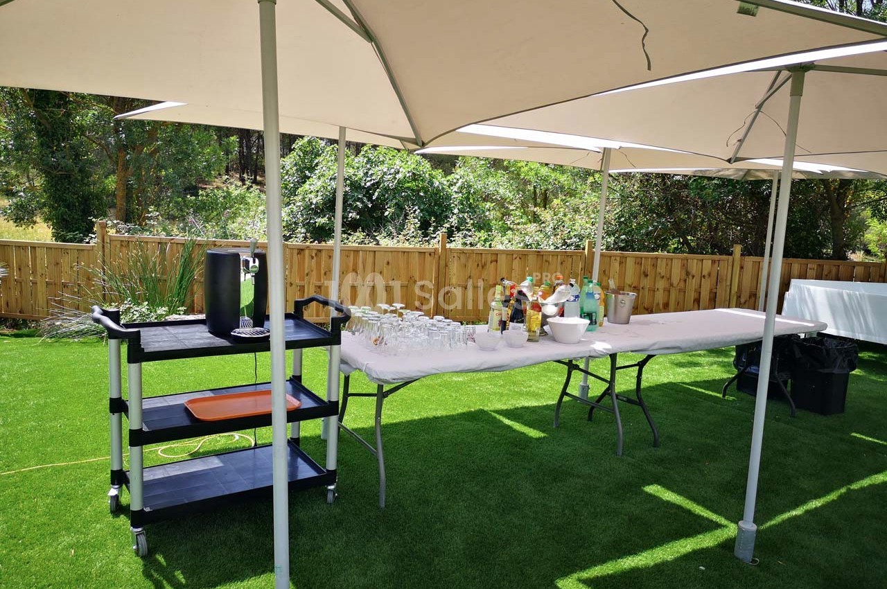 Table de buffet en extérieur avec boissons, verres et parasols, installée sur une pelouse près d'une clôture en bois.