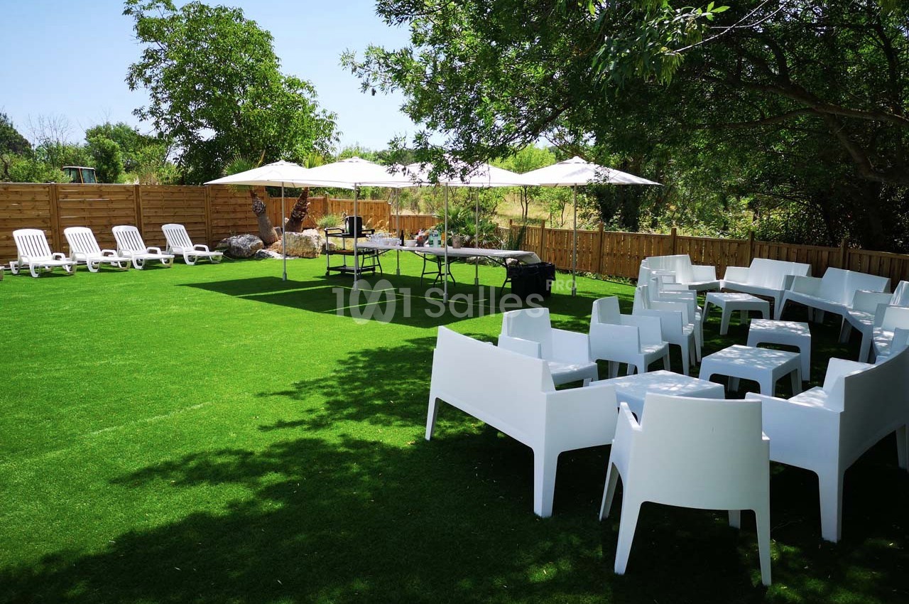 Espace extérieur avec pelouse artificielle, chaises blanches, tables et parasols sous des arbres et ciel dégagé.