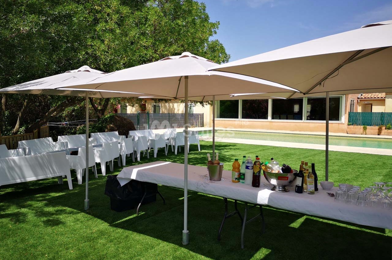 Table dressée avec boissons et verres sous des parasols blancs, entourée de chaises blanches sur une pelouse.