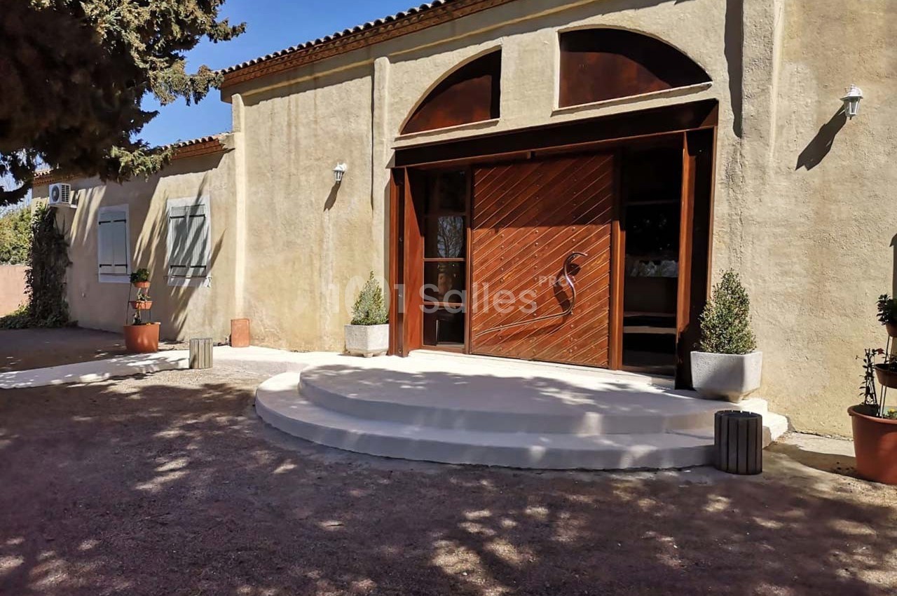 Entrée d'un bâtiment avec une porte en bois, des marches arrondies et des plantes en pots sur un sol en gravier.