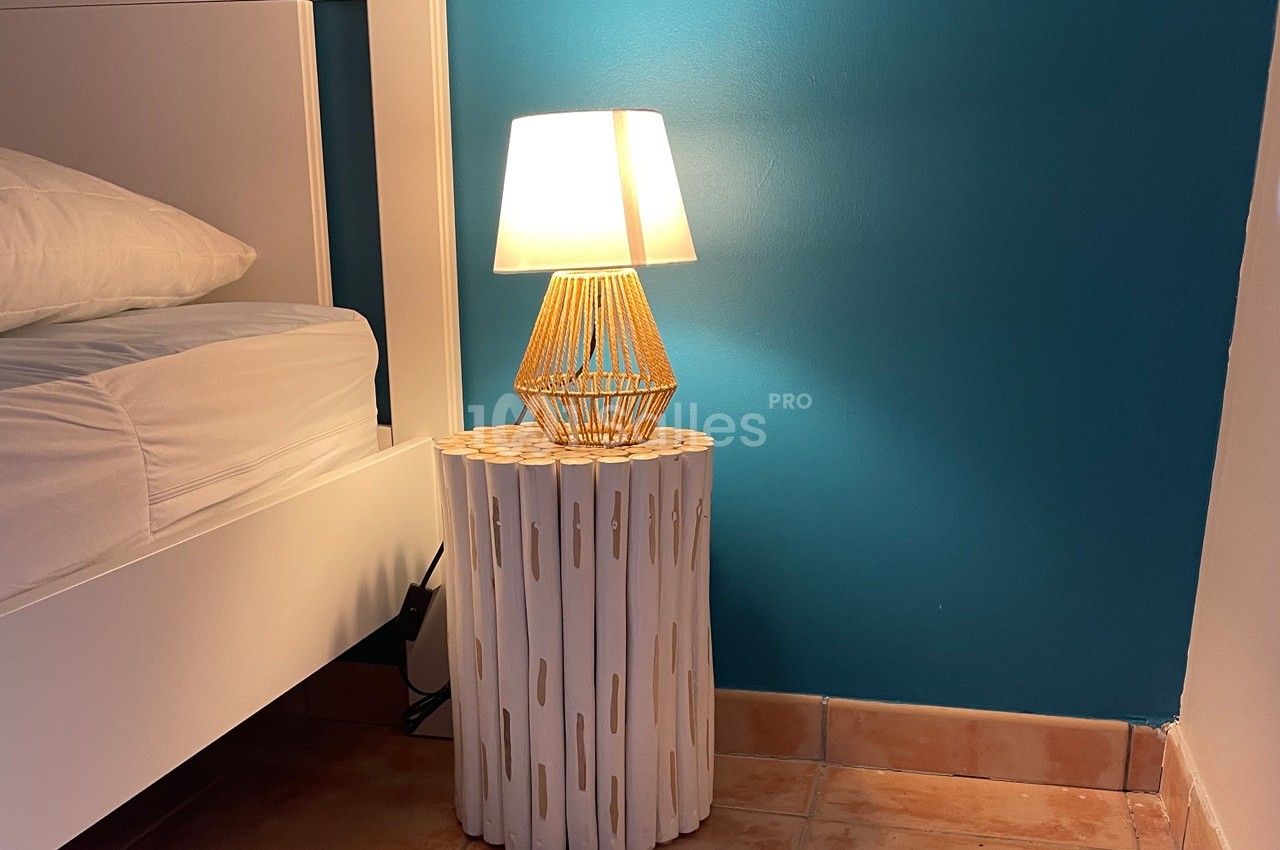 Lampe allumée sur une table de chevet en bois blanc, près d'un lit avec mur bleu en arrière-plan.