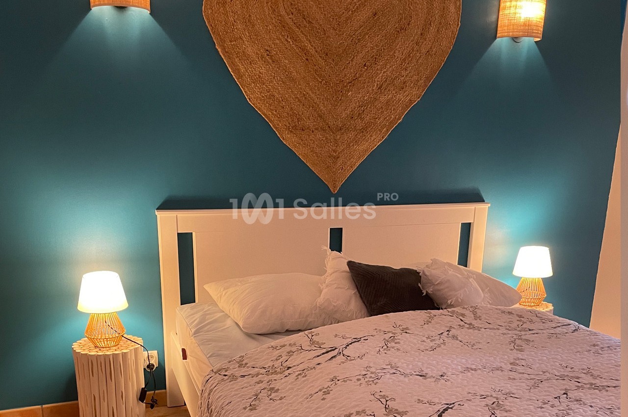 Lit double avec draps à motifs floraux, encadré par deux lampes de chevet et un mur bleu décoré d'un cœur en rotin.