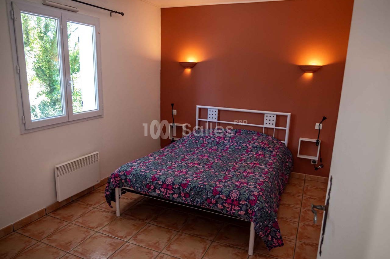 Chambre avec un lit double, mur orange, carrelage au sol, fenêtre à gauche et appliques murales allumées.