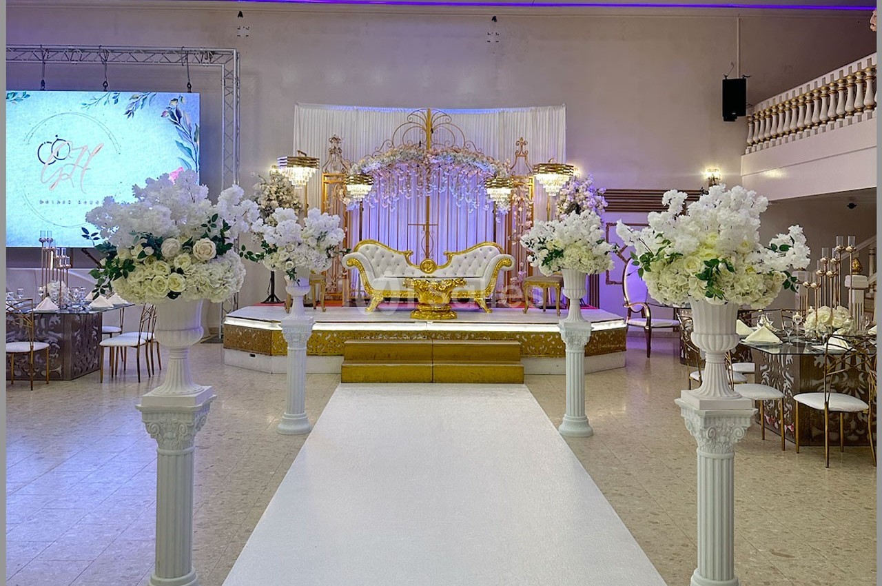 Salle de réception décorée avec une allée centrale, des colonnes fleuries et une scène ornée d'un canapé doré.