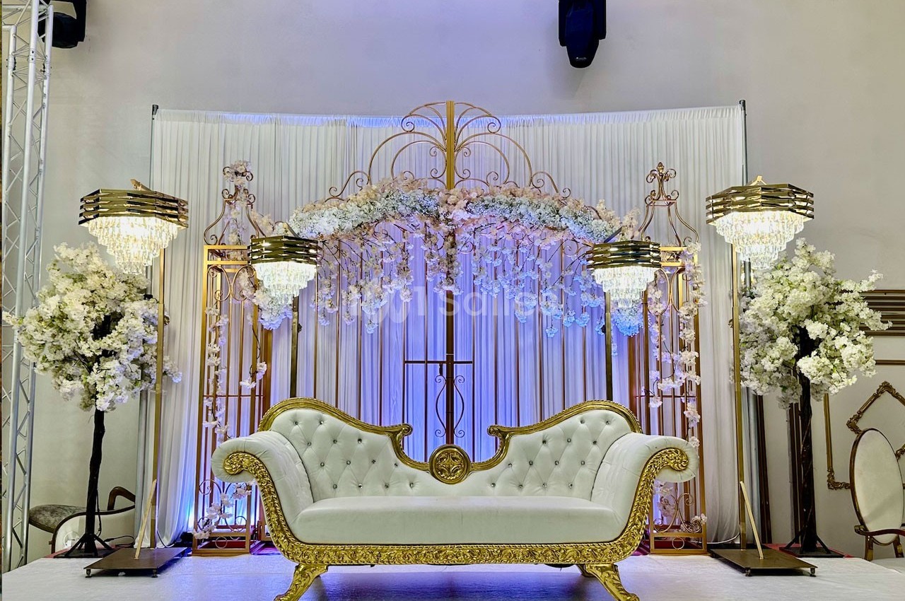 Canapé blanc et doré devant une décoration florale luxueuse avec rideaux, lustres et arches dorées.