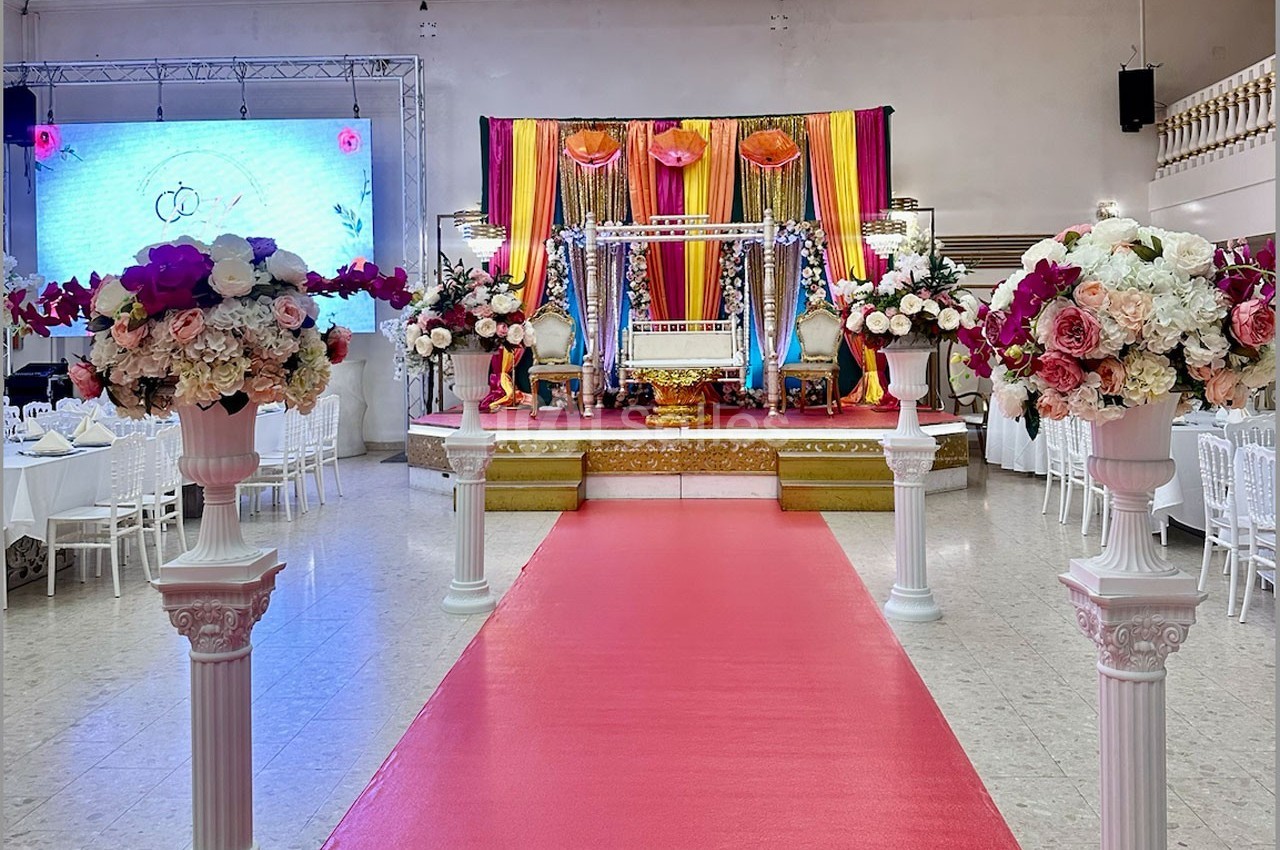 Décoration de mariage avec une allée rouge bordée de colonnes fleuries menant à une scène ornée de drapés colorés.