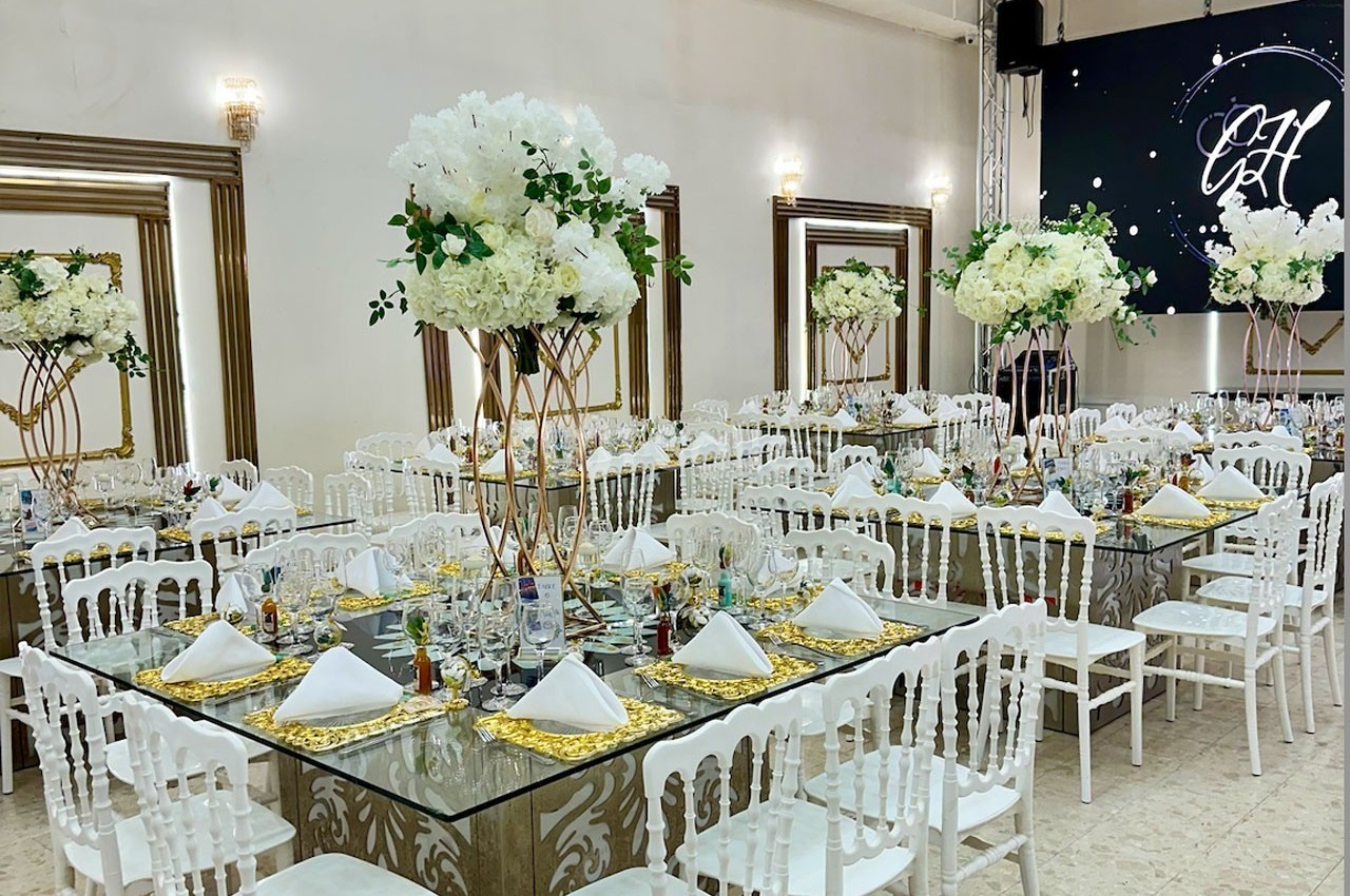 Salle de réception décorée avec des tables en verre, nappes dorées, chaises blanches et centres de table fleuris.