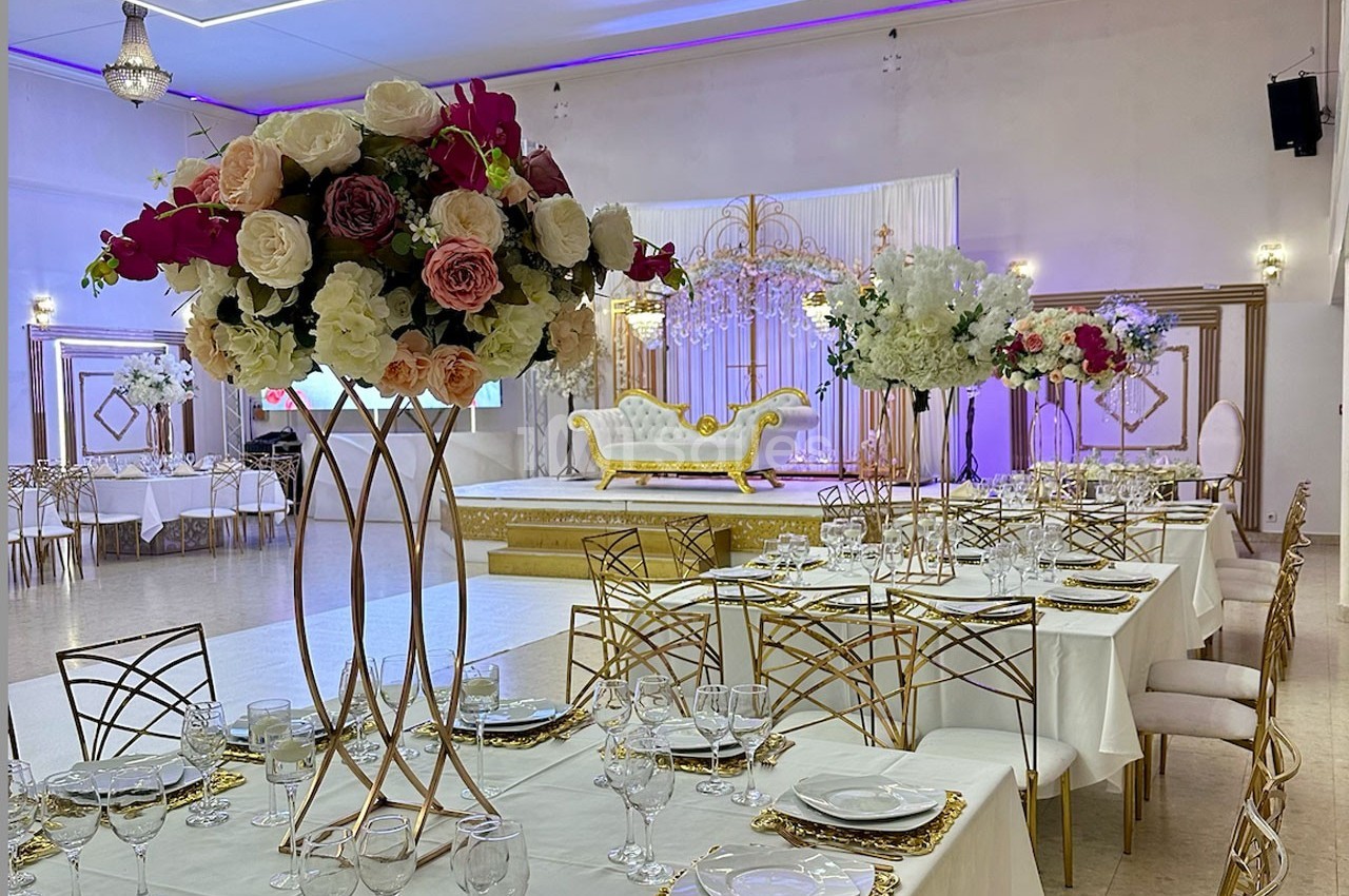 Salle de réception décorée avec des tables dressées, des centres de table fleuris et une scène ornée d'un canapé doré.