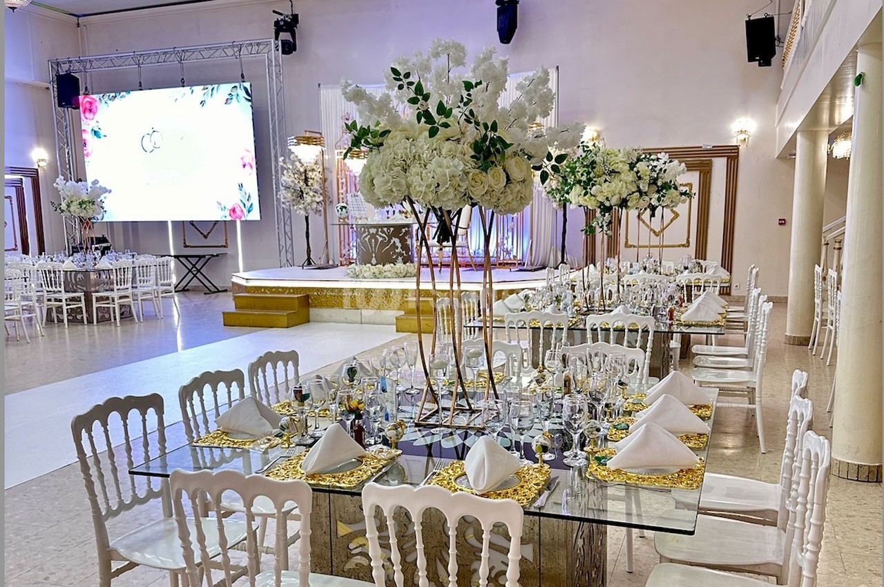 Salle de réception élégante avec tables décorées de nappes dorées, centres de table fleuris et scène en arrière-plan.