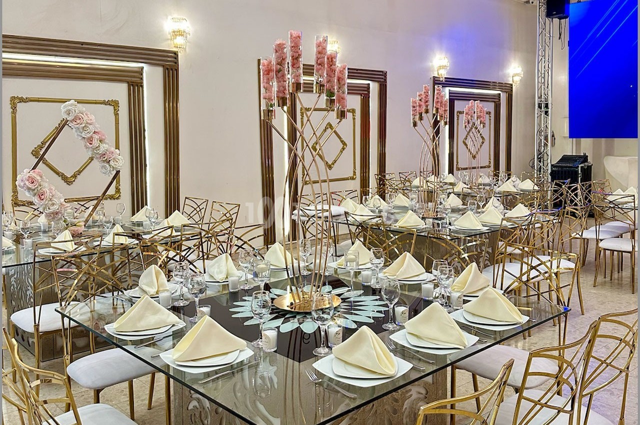 Salle de réception décorée avec des tables en verre, chaises dorées, serviettes pliées et chandeliers ornés de fleurs roses.