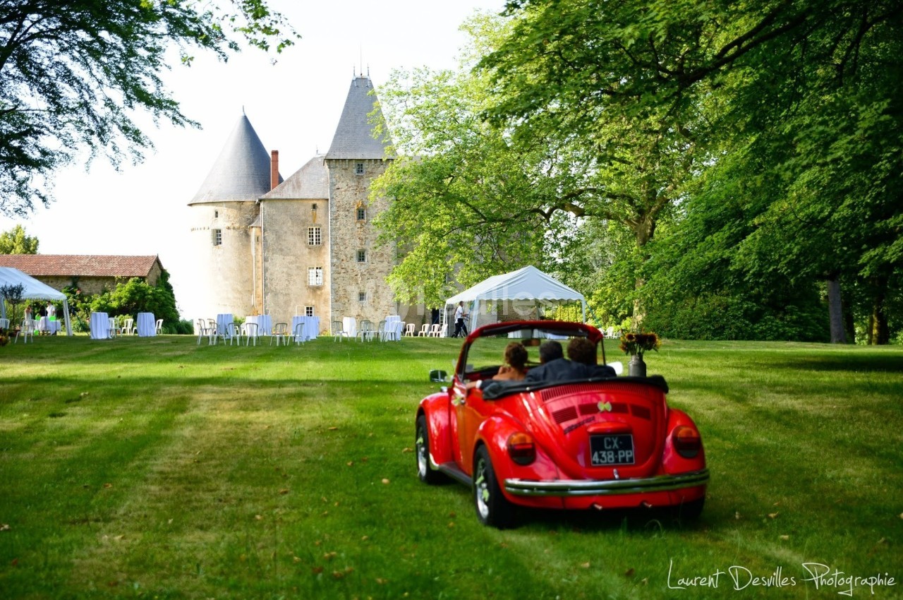 Voiture ancienne rouge décapotable dans un parc verdoyant, avec un château en arrière-plan et des tentes blanches.