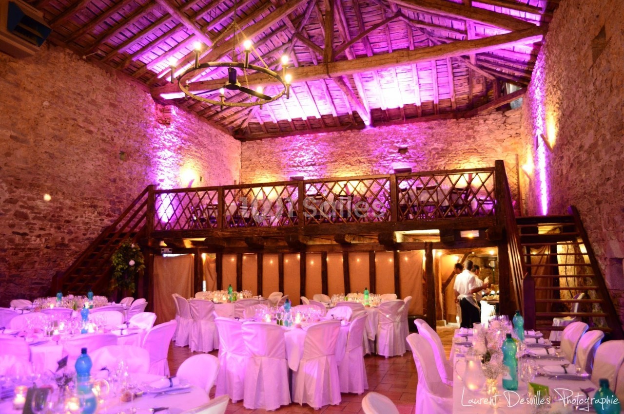 Salle de réception en pierre avec mezzanine en bois, éclairage rose et tables décorées pour un événement.