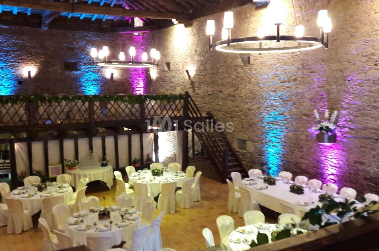 Salle de réception en pierre avec tables décorées, chaises blanches et éclairage coloré sur les murs.