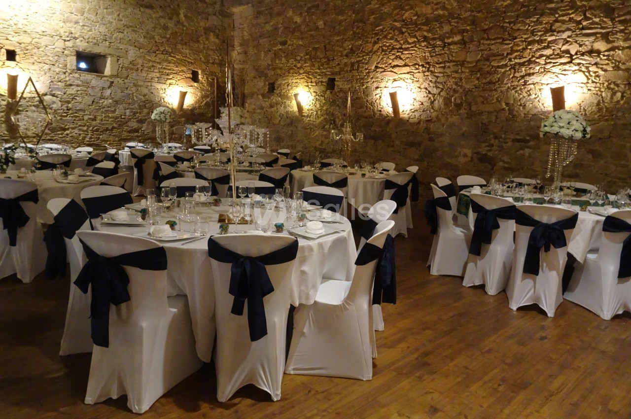 Salle de réception avec murs en pierre, tables rondes dressées, nappes blanches et chaises décorées de nœuds noirs.