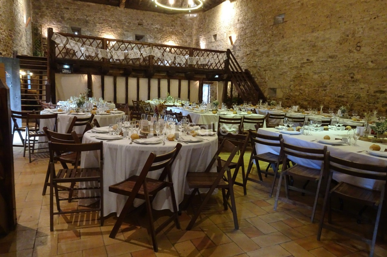 Salle de réception rustique avec tables rondes dressées, chaises en bois et mezzanine en pierre apparente.