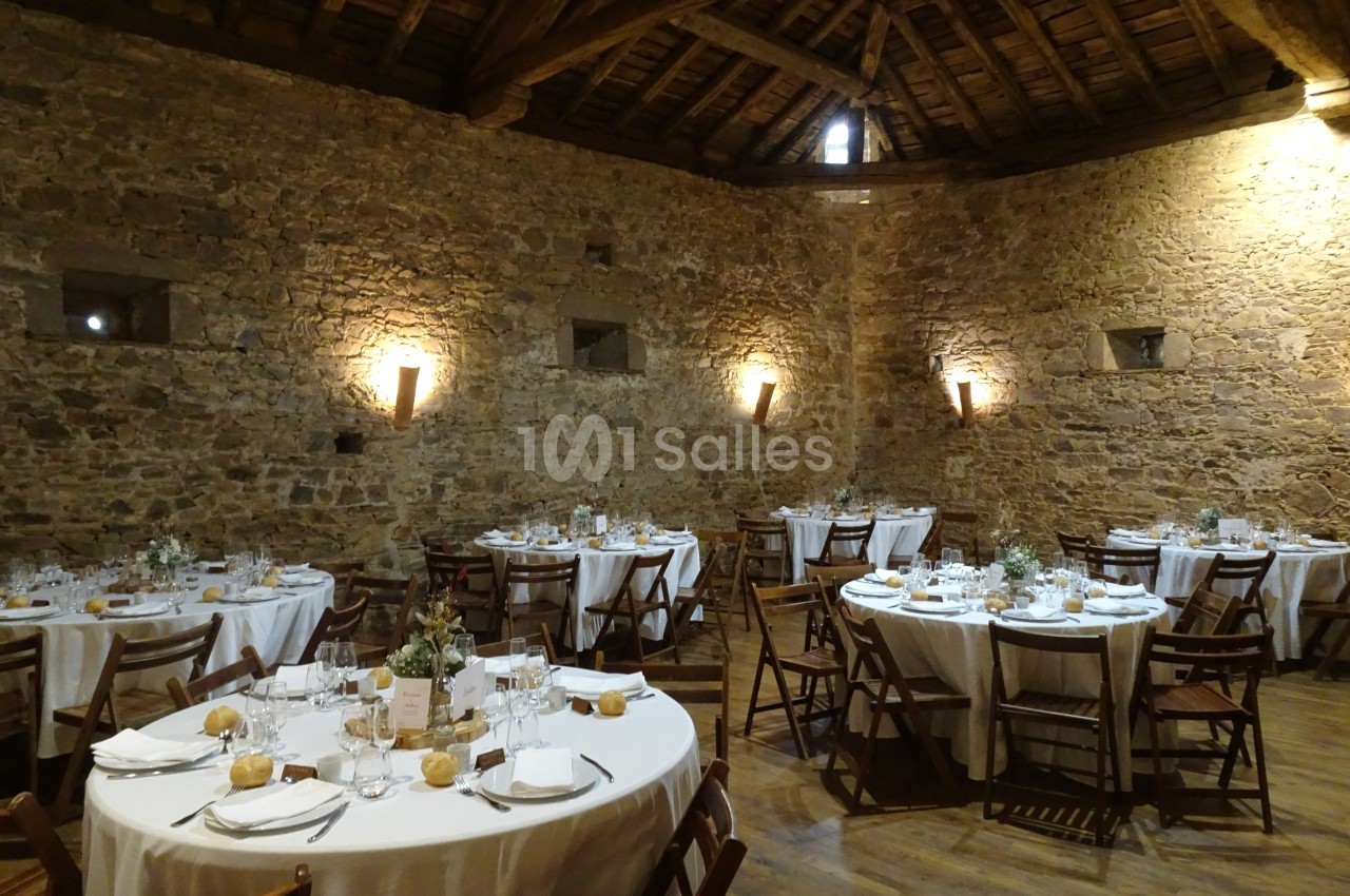 Salle en pierre avec des tables rondes dressées pour un repas, éclairée par des appliques murales.