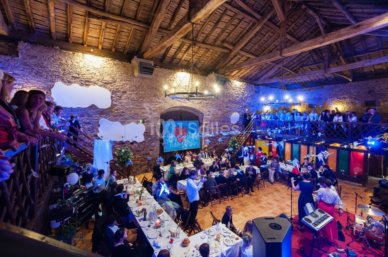 Salle en pierre avec plafond en poutres, tables dressées pour un repas, invités assis et musiciens sur scène.